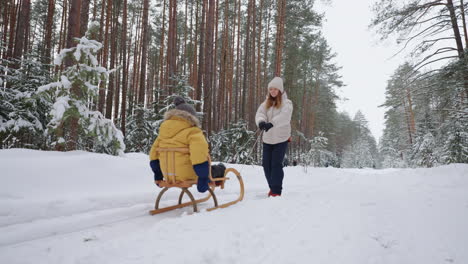 happy-young-woman-is-walking-with-her-little-child-in-winter-forest-son-is-sitting-in-wooden-sleigh